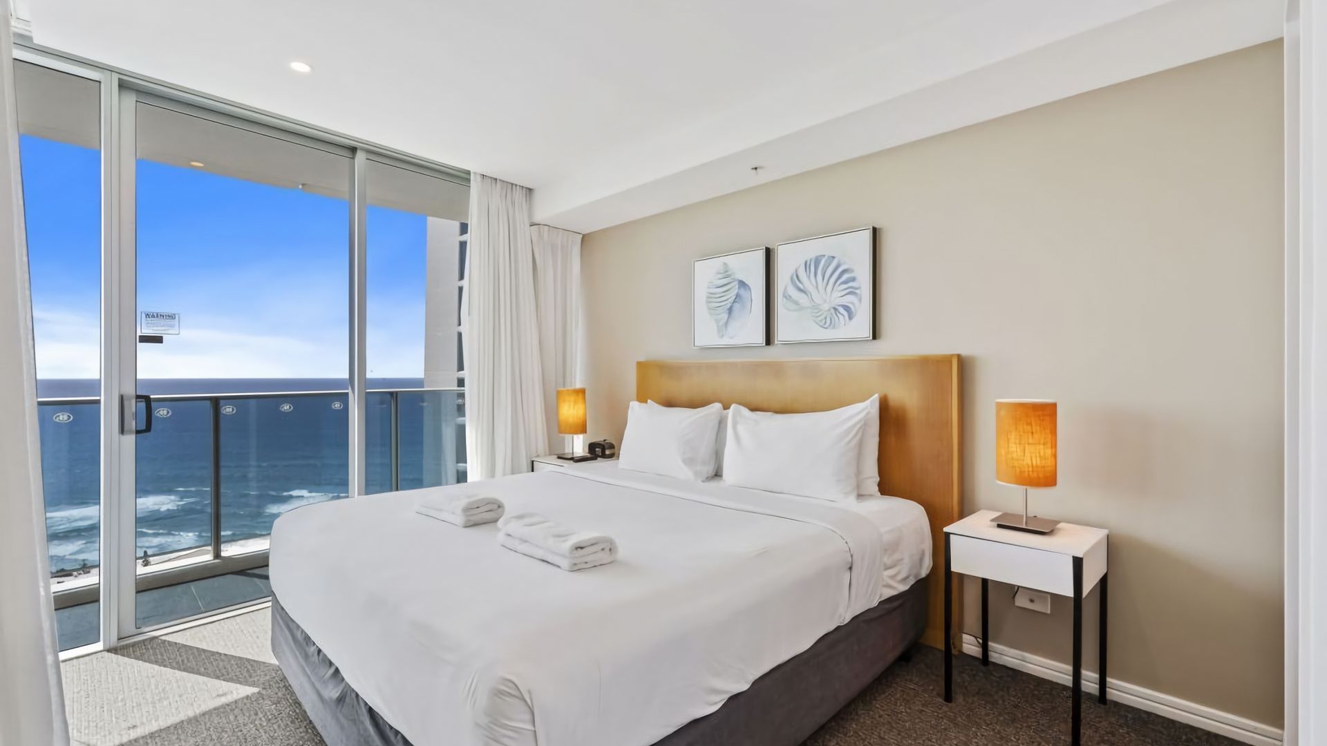 A hotel room with a large bed, two bedside lamps, a view of the ocean, and two framed artworks on the wall.