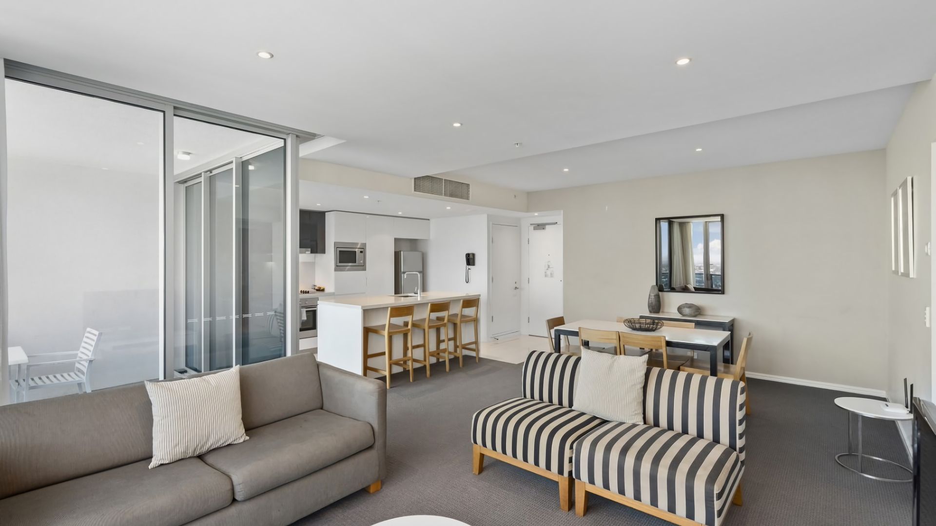 A modern living room with a sofa, a striped couch, a dining area, and a kitchen in the background.