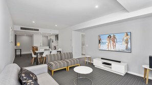 A modern living room with a sofa, a dining area, a kitchen, and a flat-screen TV mounted on the wall. - Orchid Residences Apartment 21803 (Surfers Paradise)