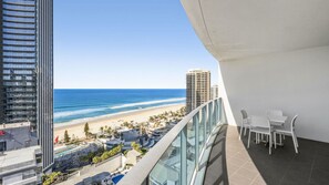 Ein Balkon mit Blick auf Strand und Meer, ein Essbereich mit Stühlen und Tisch sowie moderne Gebäude im Hintergrund.
