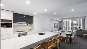 A modern kitchen with a built-in microwave, stove, and sink. There is a dining area with a table and chairs, and a living room with a sofa and a large window. - Orchid Residences Apartment 21803 (Surfers Paradise)