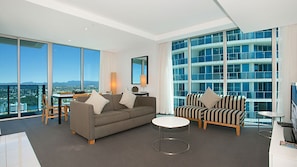A modern living room with a sofa, armchair, and coffee table, large windows, and a view of a cityscape. - Orchid Residences Apartment 22306 (Surfers Paradise)