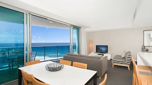 A modern living room with a large window overlooking the ocean, a dining area with a table and chairs, and a seating area with a sofa and armchairs. - Orchid Residences Apartment 22402 (Surfers Paradise)