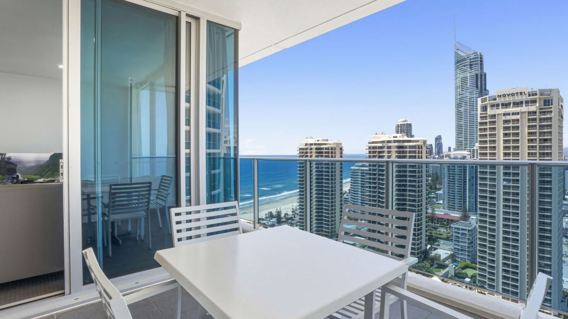 A balcony with a white table and chairs, overlooking a cityscape and the ocean.