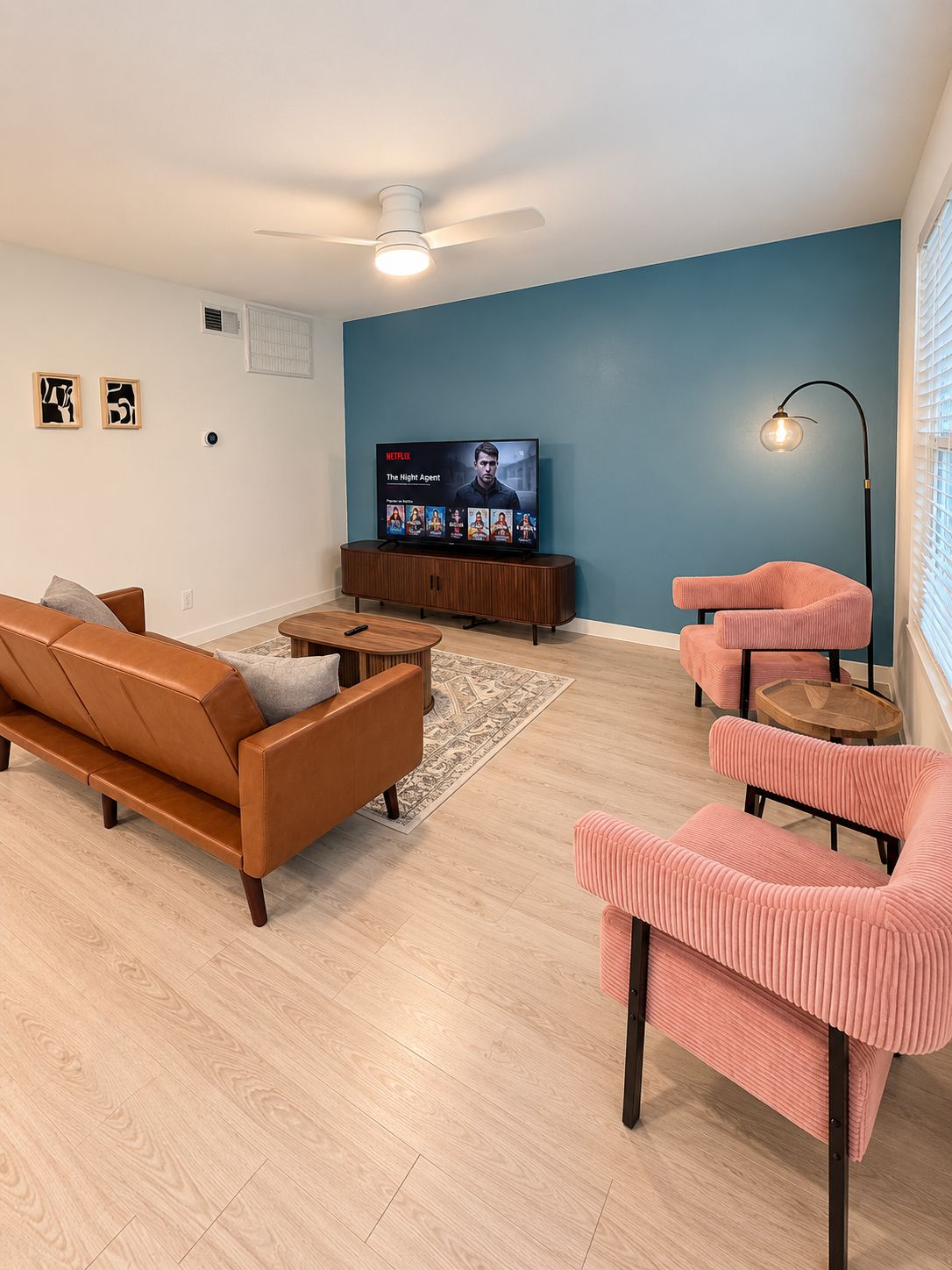 A living room with a brown leather sofa, a pink armchair, a wooden coffee table, a TV on a stand, and a ceiling fan.