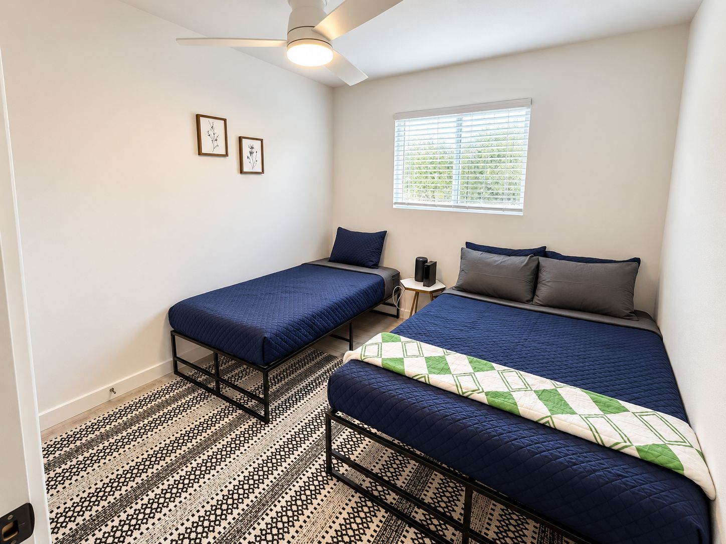 A bedroom with two single beds, a large bed, a ceiling fan, a window with blinds, and a patterned rug.