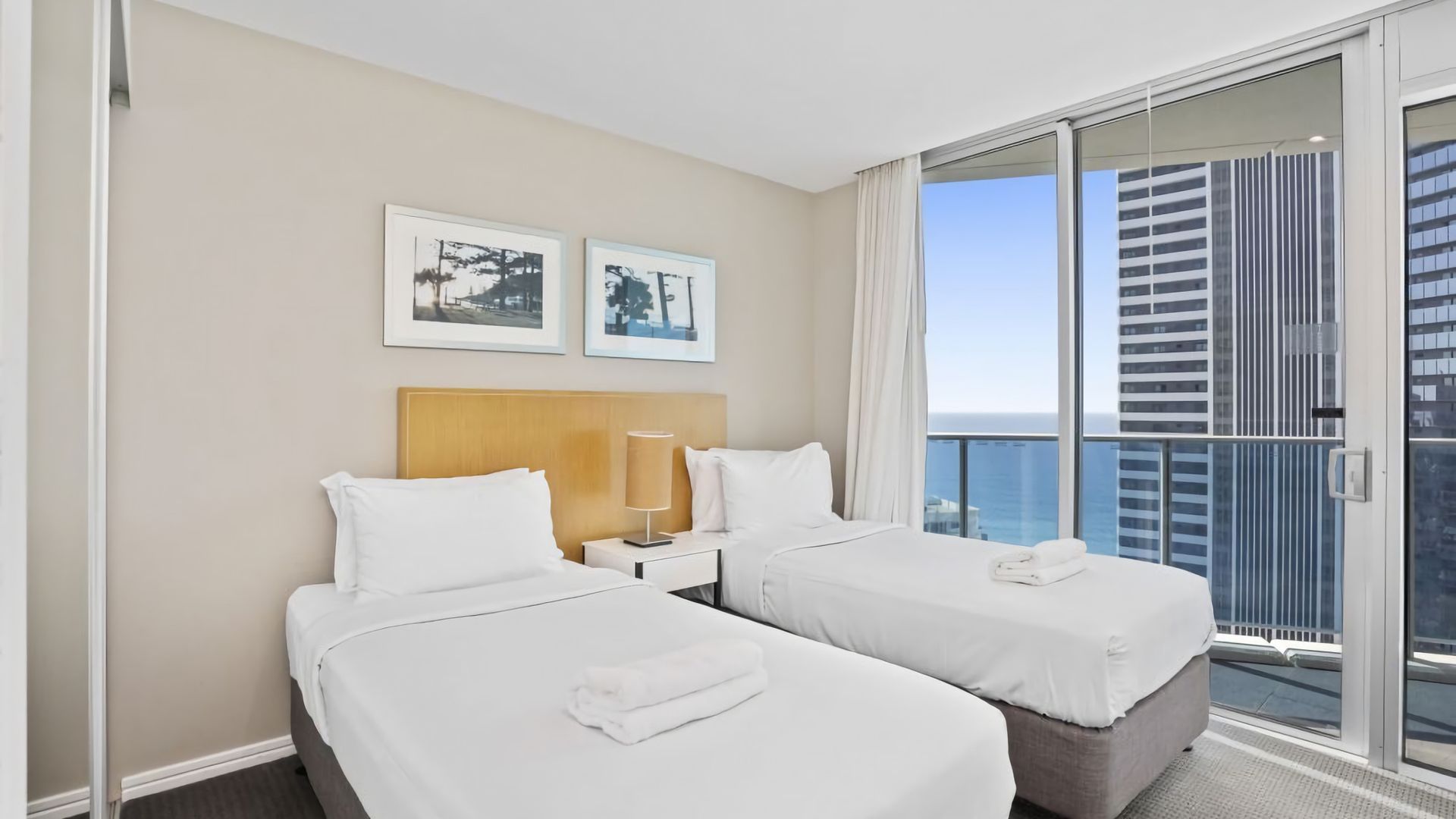 A hotel room with two beds, a nightstand, a lamp, and a view of the city.