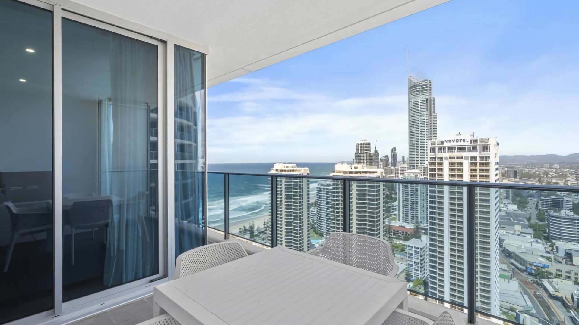 A balcony with a table and chairs, overlooking a cityscape and the ocean.