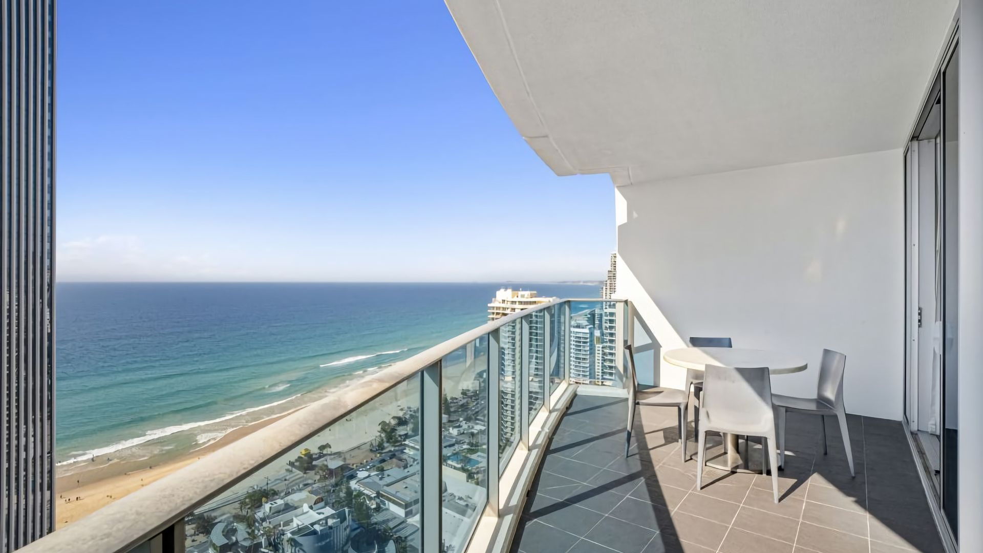 A balcony with a view of the ocean and beach, a table with chairs, and a glass railing.