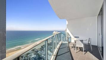 A balcony with a view of the ocean and beach, a table with chairs, and a glass railing.