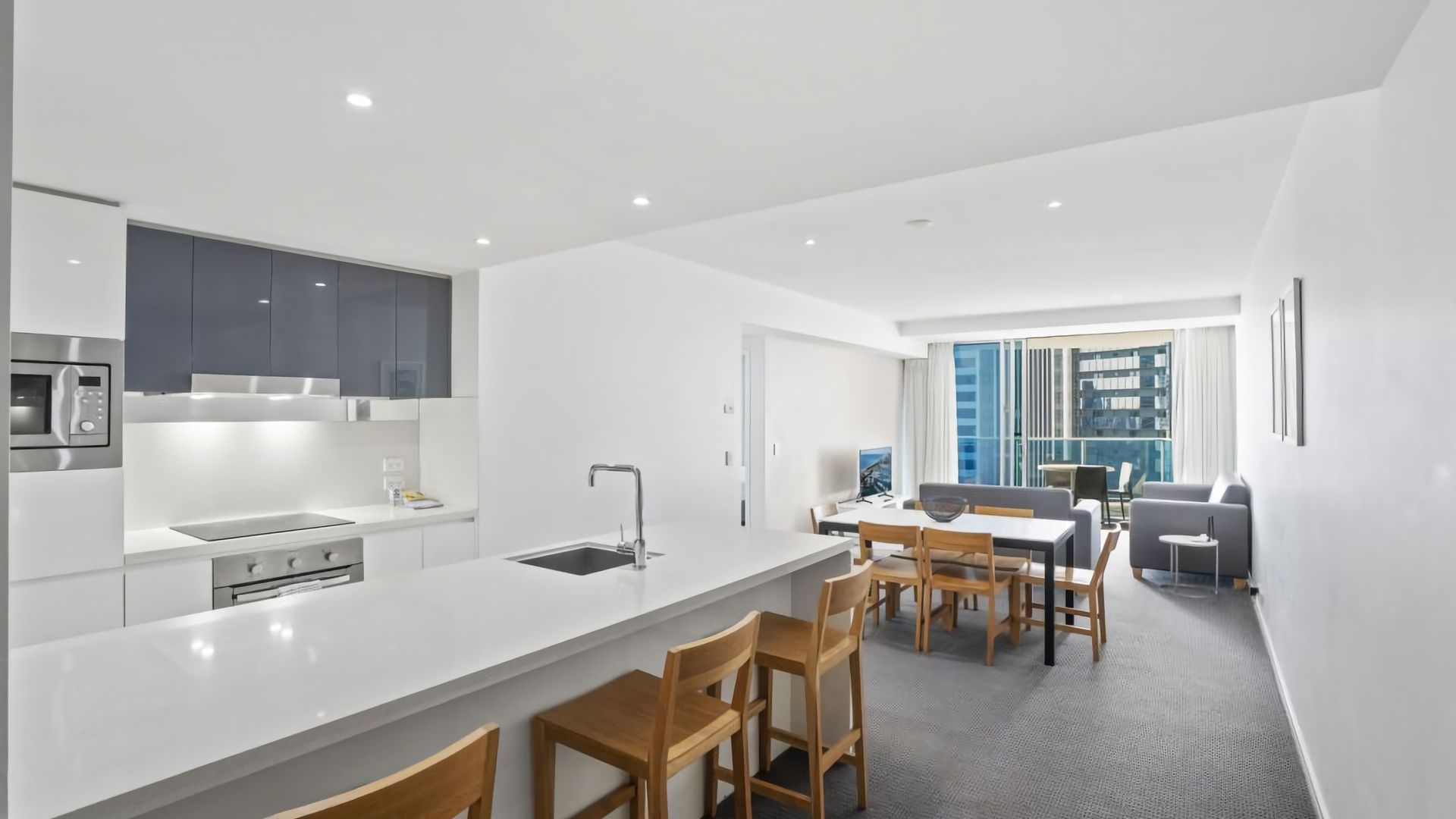 A modern kitchen with a central island, dining area with wooden chairs, and a view of a cityscape through large windows.
