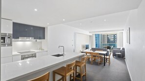 A modern kitchen with a central island, dining area with wooden chairs, and a view of a cityscape through large windows. - Orchid Residences Apartment 23003 (Surfers Paradise)
