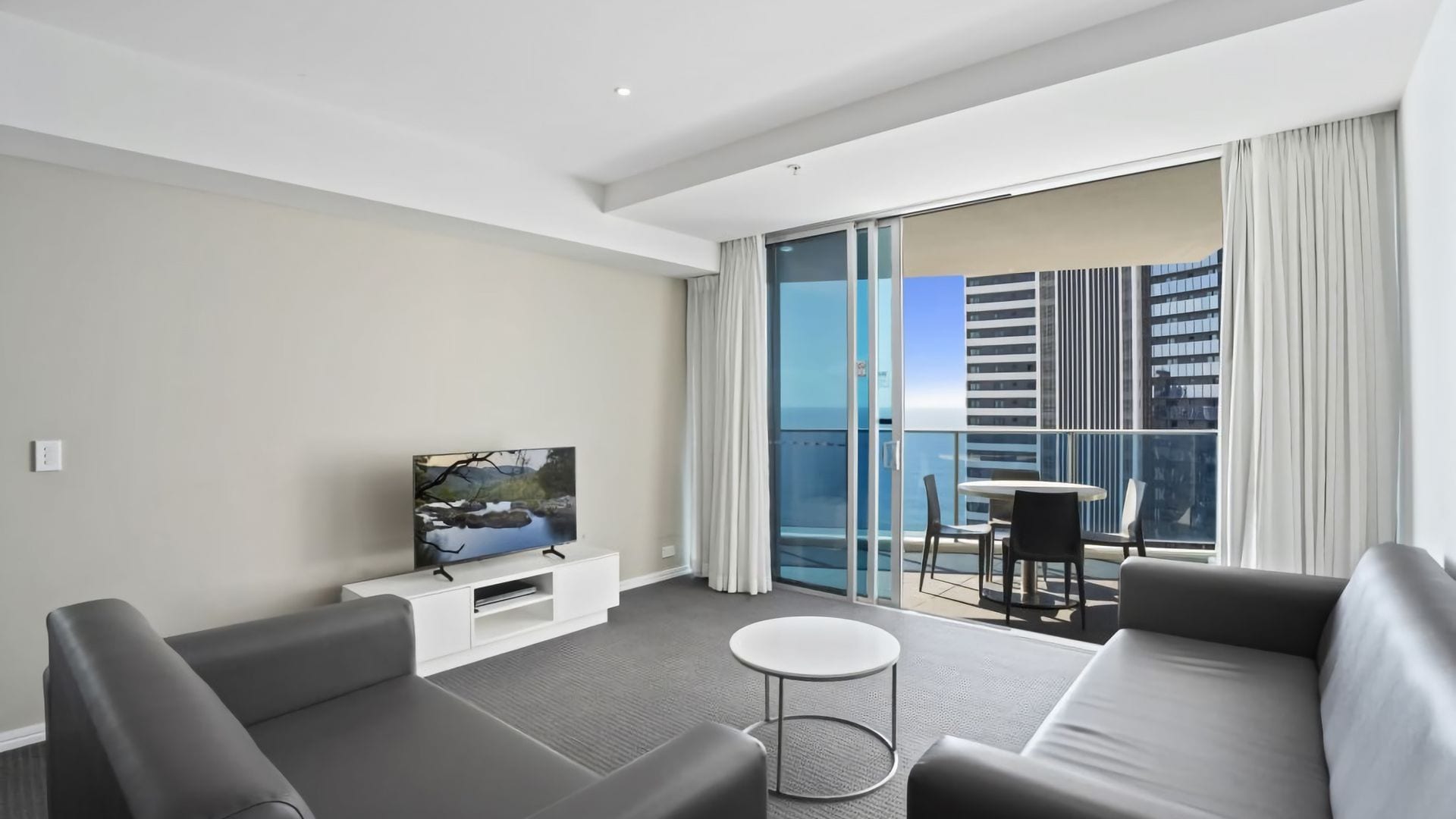 A modern hotel room with a sofa, a small table, a TV on a stand, and a balcony with a city view.