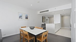A modern kitchen with white cabinetry, a central island, and a dining area with a white table and wooden chairs. - Orchid Residences Apartment 23003 (Surfers Paradise)