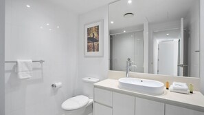 A modern bathroom with a white sink, toilet, and shower. There is a framed picture on the wall and a large mirror above the sink. - Orchid Residences Apartment 23003 (Surfers Paradise)