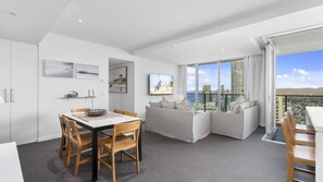 A modern living room with a dining area, a sofa set, and a large window offering a city view. - Orchid Residences Apartment 23405 (Surfers Paradise)