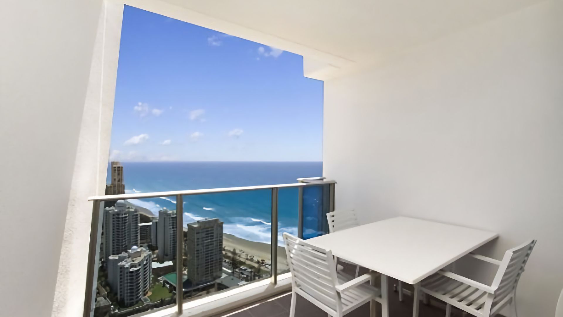A balcony with a white table and chairs, overlooking a cityscape and the ocean.