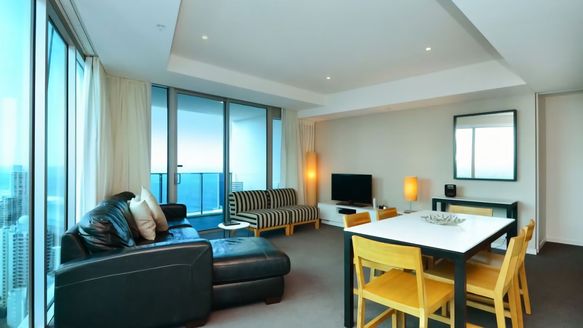 A modern hotel room with a large window, a sofa, a dining table, and a TV.