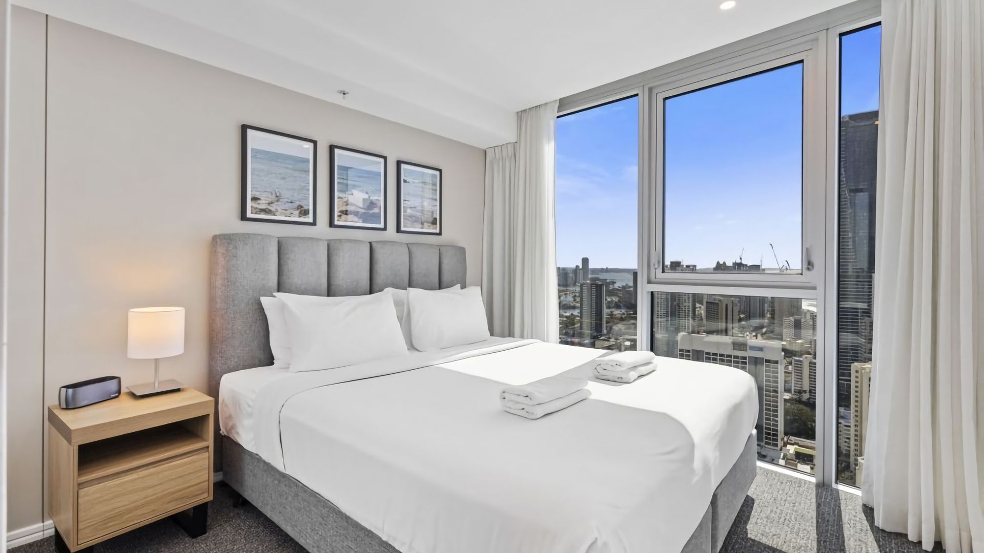 A hotel room with a large bed, a nightstand, a lamp, and a city view through large windows.