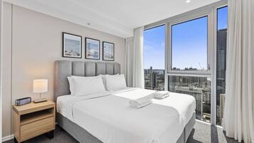 A hotel room with a large bed, a nightstand, a lamp, and a city view through large windows.