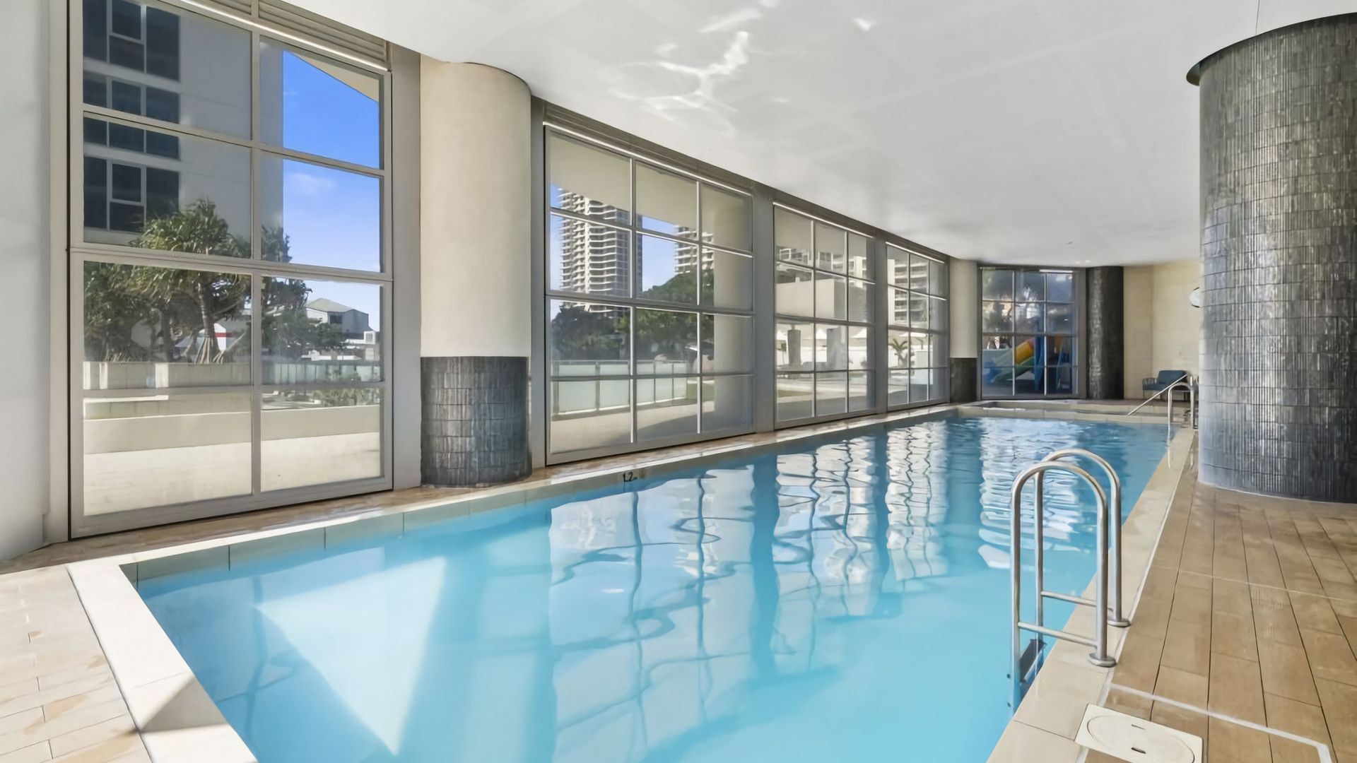 An indoor swimming pool with clear glass walls, a tiled floor, and a metal ladder.