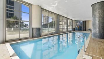 An indoor swimming pool with clear glass walls, a tiled floor, and a metal ladder.