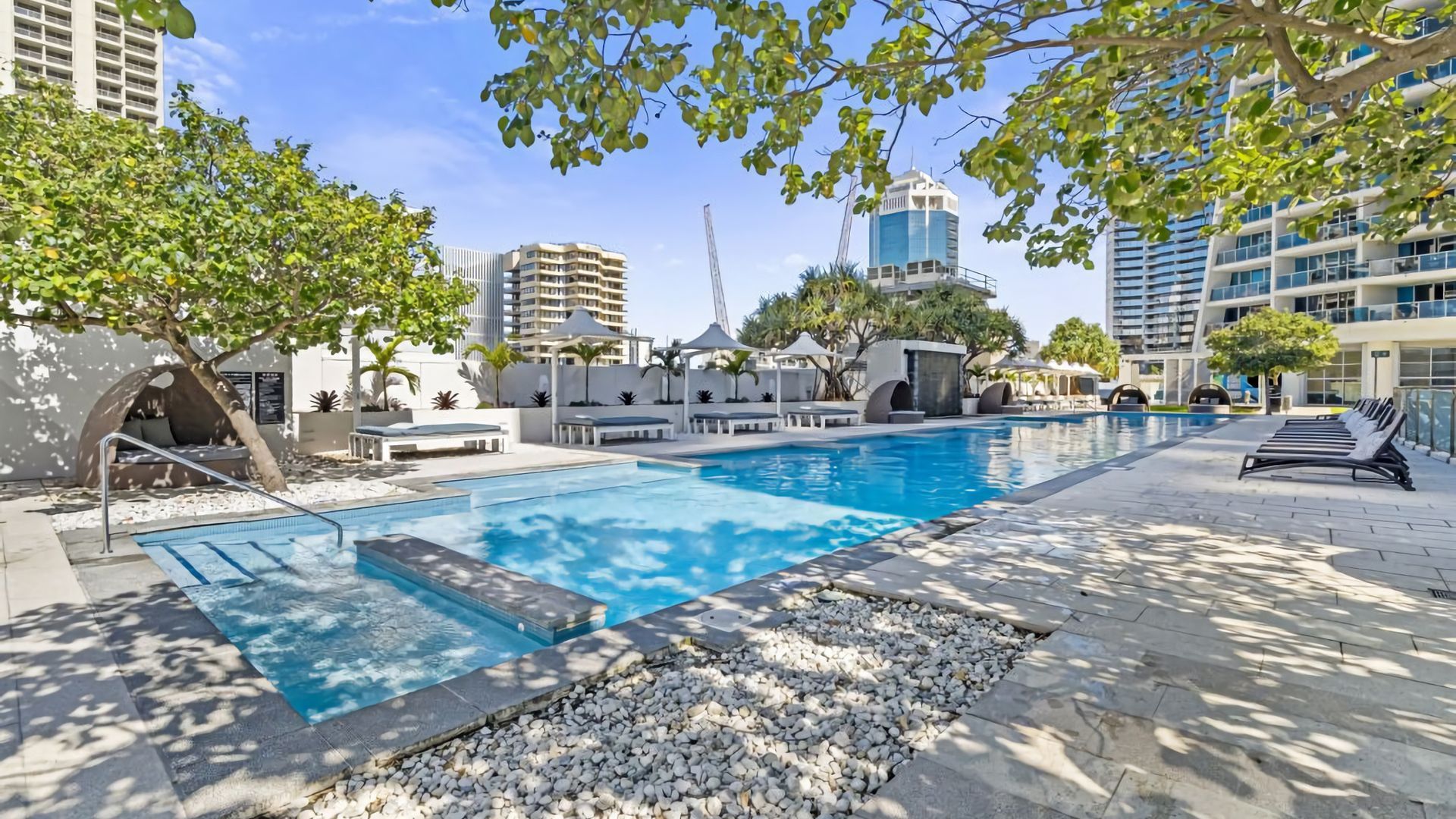 A swimming pool with lounge chairs and umbrellas, surrounded by trees and modern buildings.