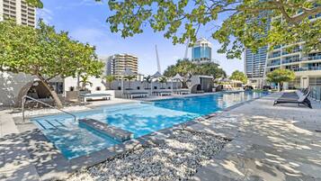 A swimming pool with lounge chairs and umbrellas, surrounded by trees and modern buildings.