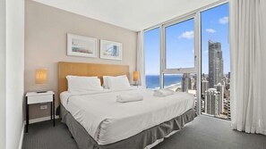 A hotel room with a large bed, two bedside lamps, a nightstand, and a view of the city and ocean. - Orchid Residences Apartment 23805 (Surfers Paradise)