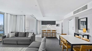 A modern hotel room with a sofa, a striped couch, a dining table with chairs, a kitchen area, and a large window offering a view. - Orchid Residences Apartment 23805 (Surfers Paradise)