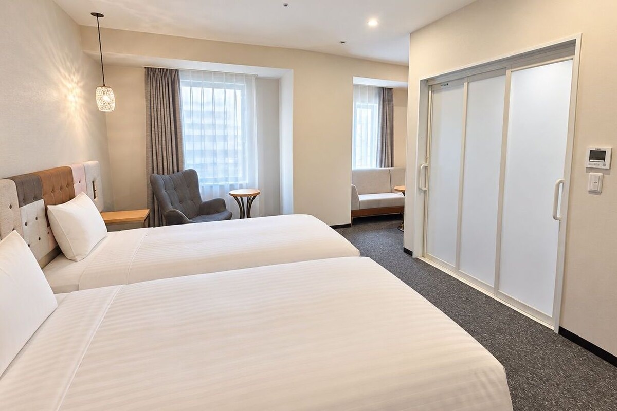 A hotel room with two beds, a chair, a small table, and a wardrobe.