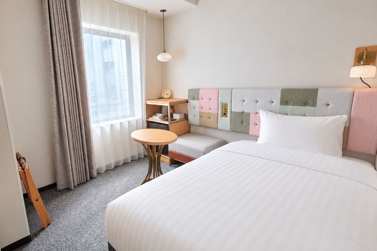 A hotel room with a bed, a small round table, a television, a window with curtains, and a headboard with multiple pillows.