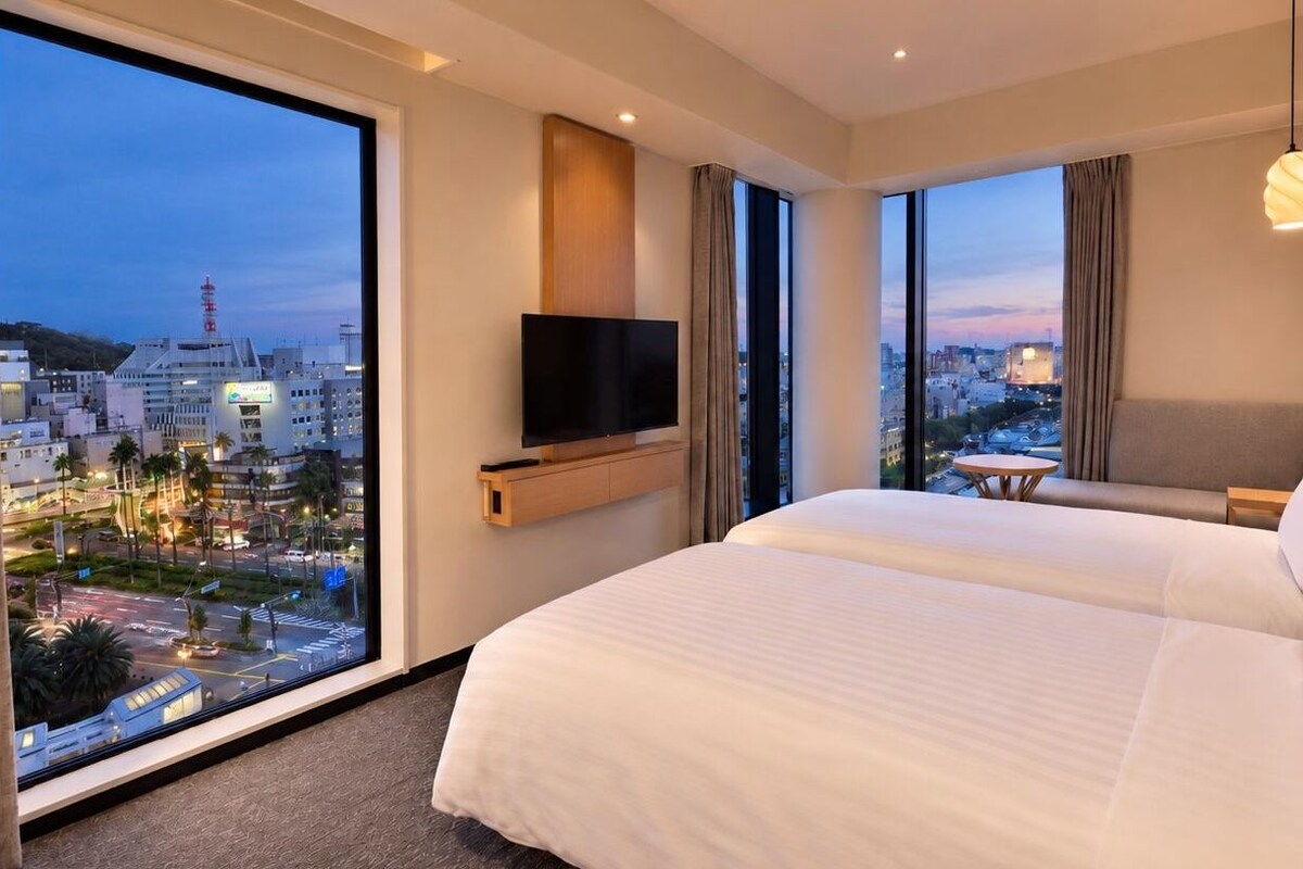 A hotel room with a large bed, a flat-screen TV, a city view, and a small table.