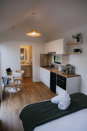 Habitación de hotel compacta con kitchenette, cama con cubrecama verde, zona de comedor con mesa y sillas, y lámpara colgante.