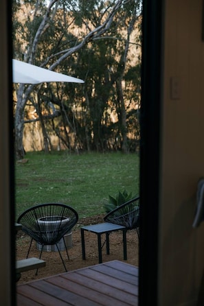 An outdoor patio with a wooden table and two chairs, a white umbrella, and a view of a grassy area with trees.