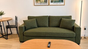 Un sofà verd amb un comandament sobre una taula de centre de fusta, una taula rodona petita amb una planta en test i dos quadres al mur.