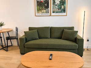 Un canapé vert avec une télécommande posée sur une table basse en bois, une petite table ronde sur laquelle se trouve un pot de plante, et deux tableaux encadrés accrochés au mur.