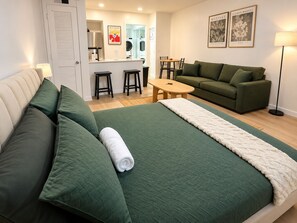 A hotel room with a bed, a green sofa, a small table, and a kitchenette in the background.