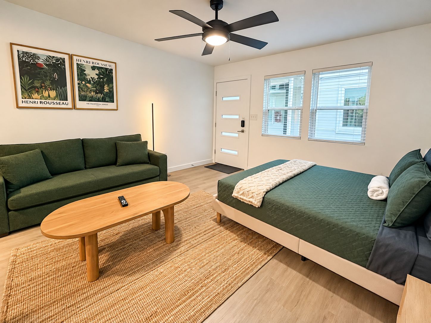 Een kamer met een groene bank, een houten tafel, een bed met een groene sprei en twee ingelijste posters aan de muur.