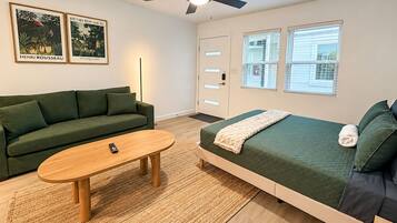 Een kamer met een groene bank, een houten tafel, een bed met een groene sprei en twee ingelijste posters aan de muur.
