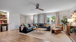 A modern living room with a black leather sofa, a wooden coffee table, and a wooden armchair. There is a ceiling fan, a bookshelf, and a potted plant. - Gilded Renegade Retreat - Walk to Doak, Sleeps 10 (Tallahassee)