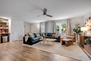 Een moderne woonkamer met een zwarte leren bank, een houten salontafel en een houten fauteuil. Er is een plafondventilator, een boekenkast en een kamerplant.