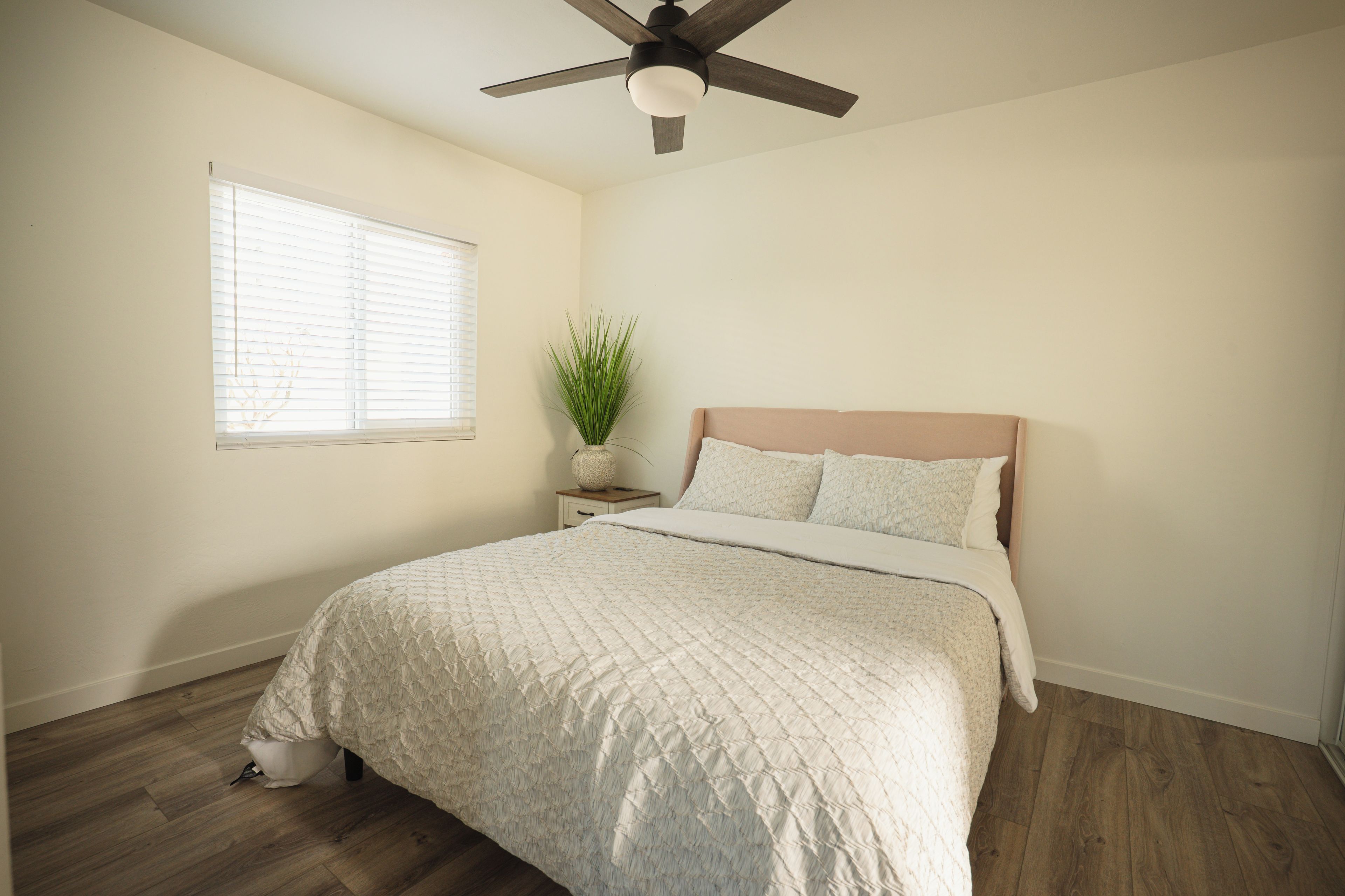 Un dormitorio con cama, ventilador de techo, ventana con persianas, planta en la mesita de noche y suelo de madera.