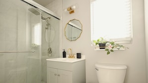 Ein modernes Badezimmer mit Duschkabine aus Glas, einer weißen Toilette, einem Waschtisch mit goldfarbenem Wasserhahn und einem Spiegel mit goldener Rahmung.