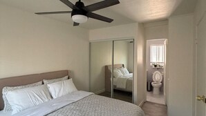 Ein Schlafzimmer mit einem Bett, einem Deckenventilator, einem Spiegel und Blick ins Bad.