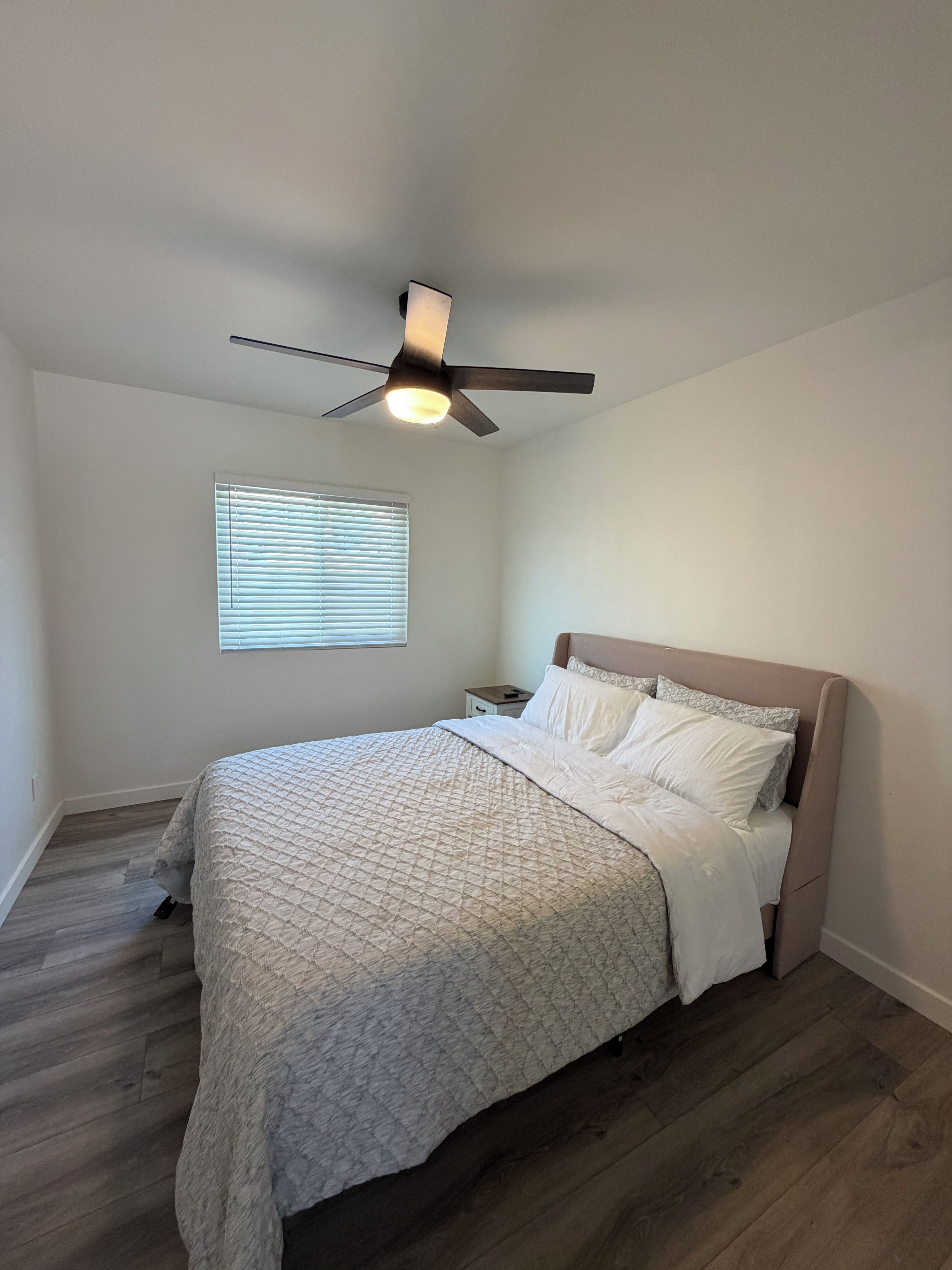 Un dormitorio con cama, mesita de noche, ventilador de techo y ventana con persianas.