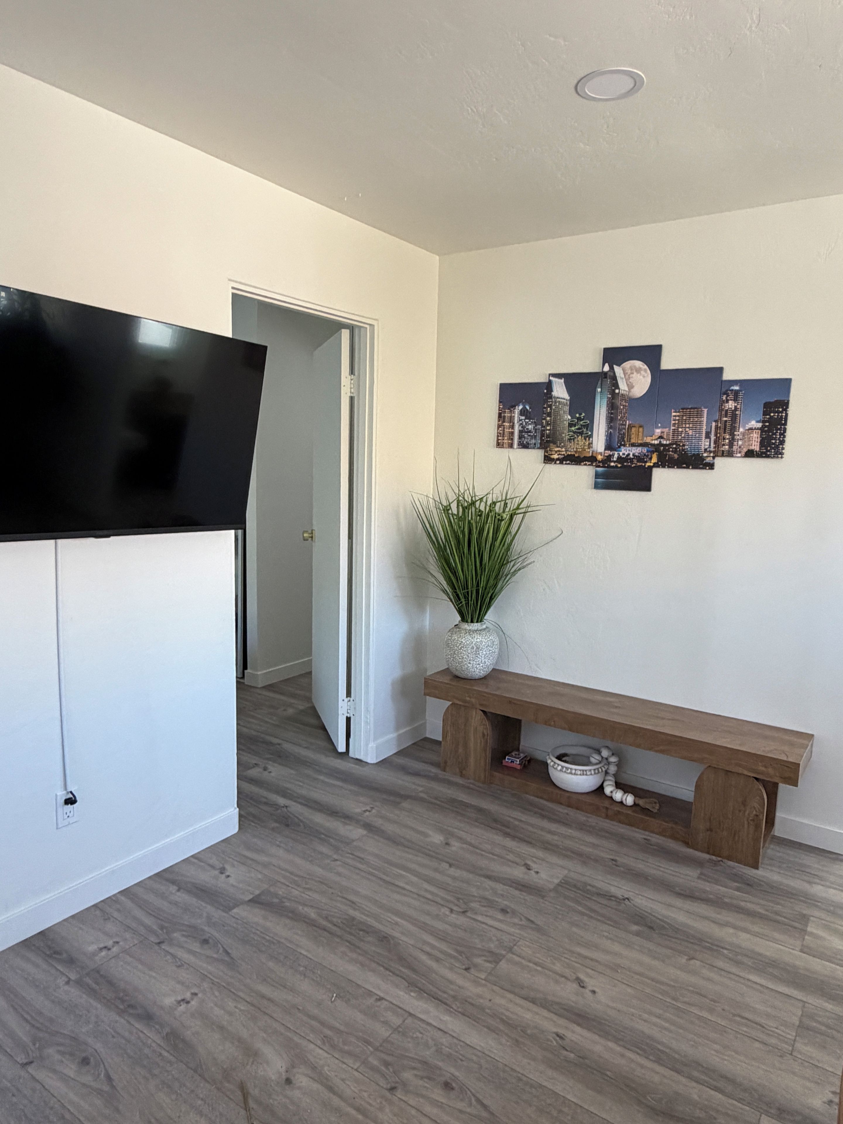 Una habitación con una televisión de pantalla plana montada en la pared, un banco de madera con un jarrón y objetos decorativos, y una obra de arte con un paisaje urbano en la pared.