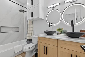 Ein modernes Badezimmer mit Badewanne, Toilette, Waschtisch mit zwei Becken und einem Spiegel mit integrierter Beleuchtung.