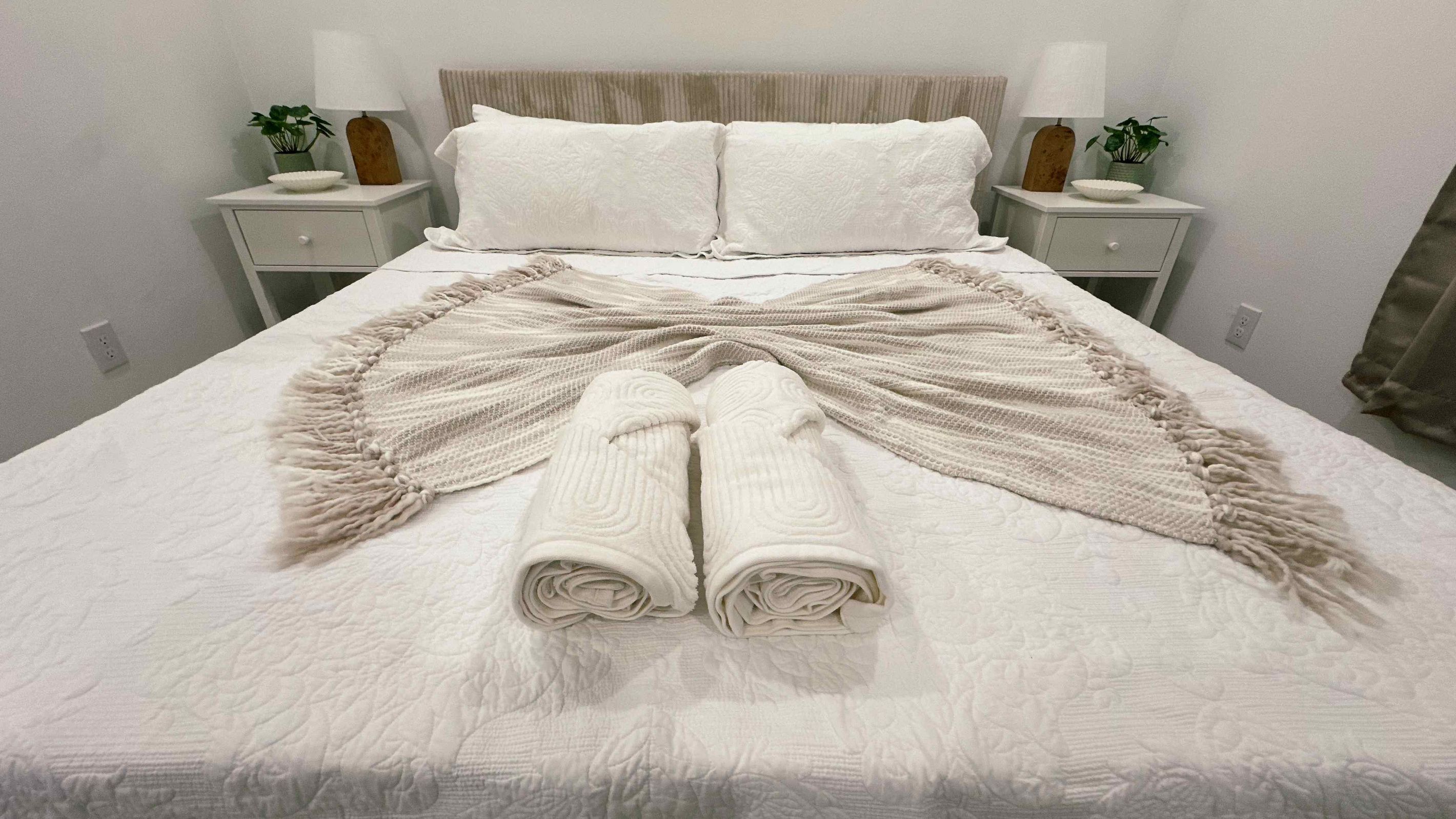 Una cama bien hecha con edredón blanco, dos toallas enrolladas y una manta decorativa.