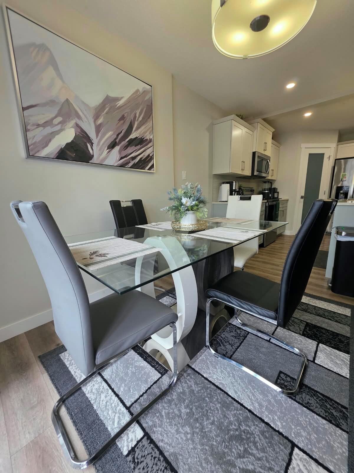 Un comedor moderno con una mesa de vidrio, sillas negras y blancas, un centro de mesa decorativo y una cocina al fondo.